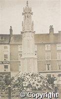 Postcard: black and white photograph of unidentified war memorial with flowers at the base and a small group of onlookers