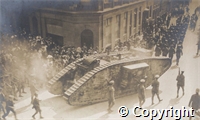 Postcard: black and white photograph of tank and troops parading along a street with crowds of onlookers